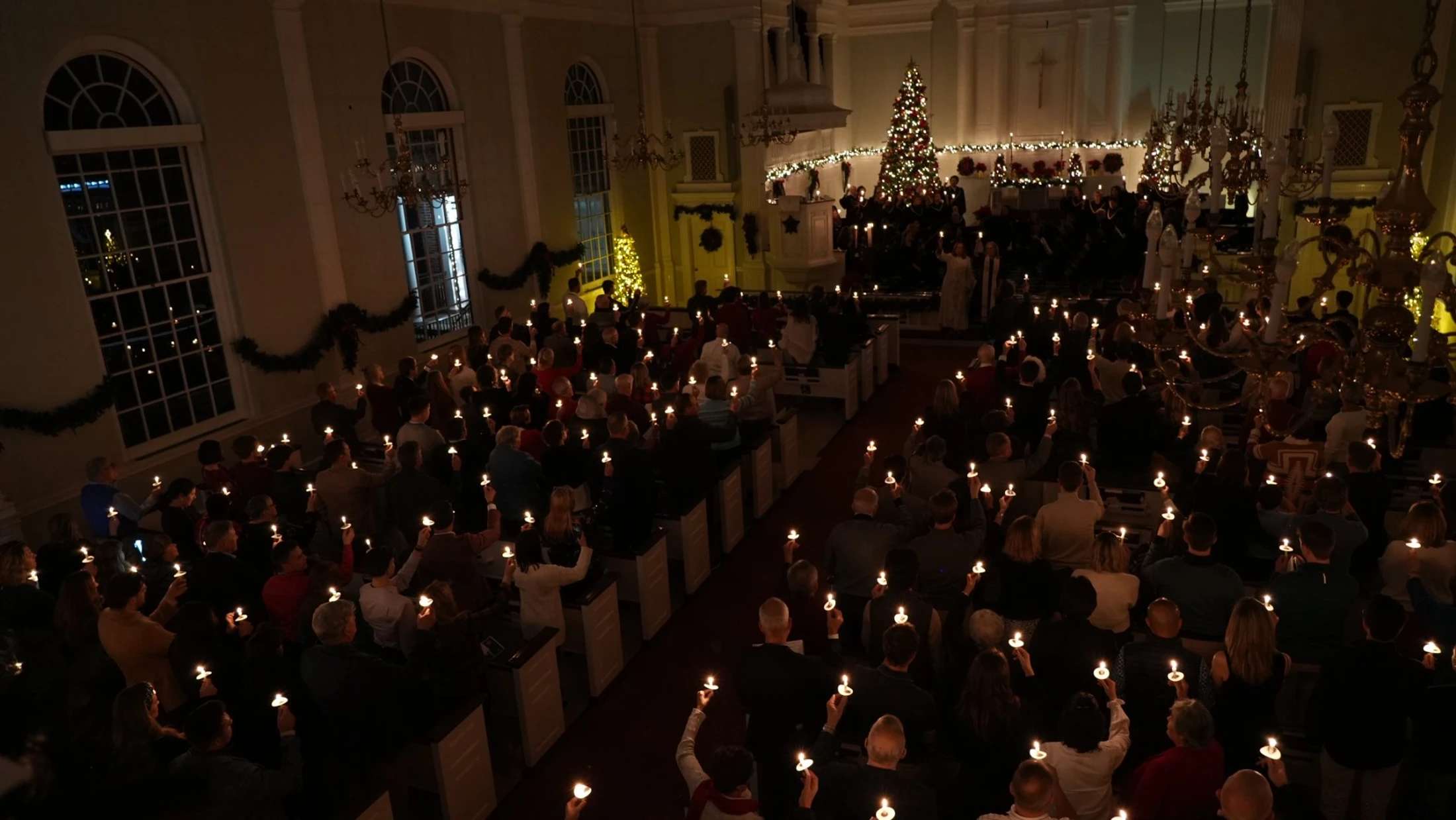 High-angle view of a congregation holding lit candles in a dimly lit church during a Christmas service, with a choir and decorated tree at the front.